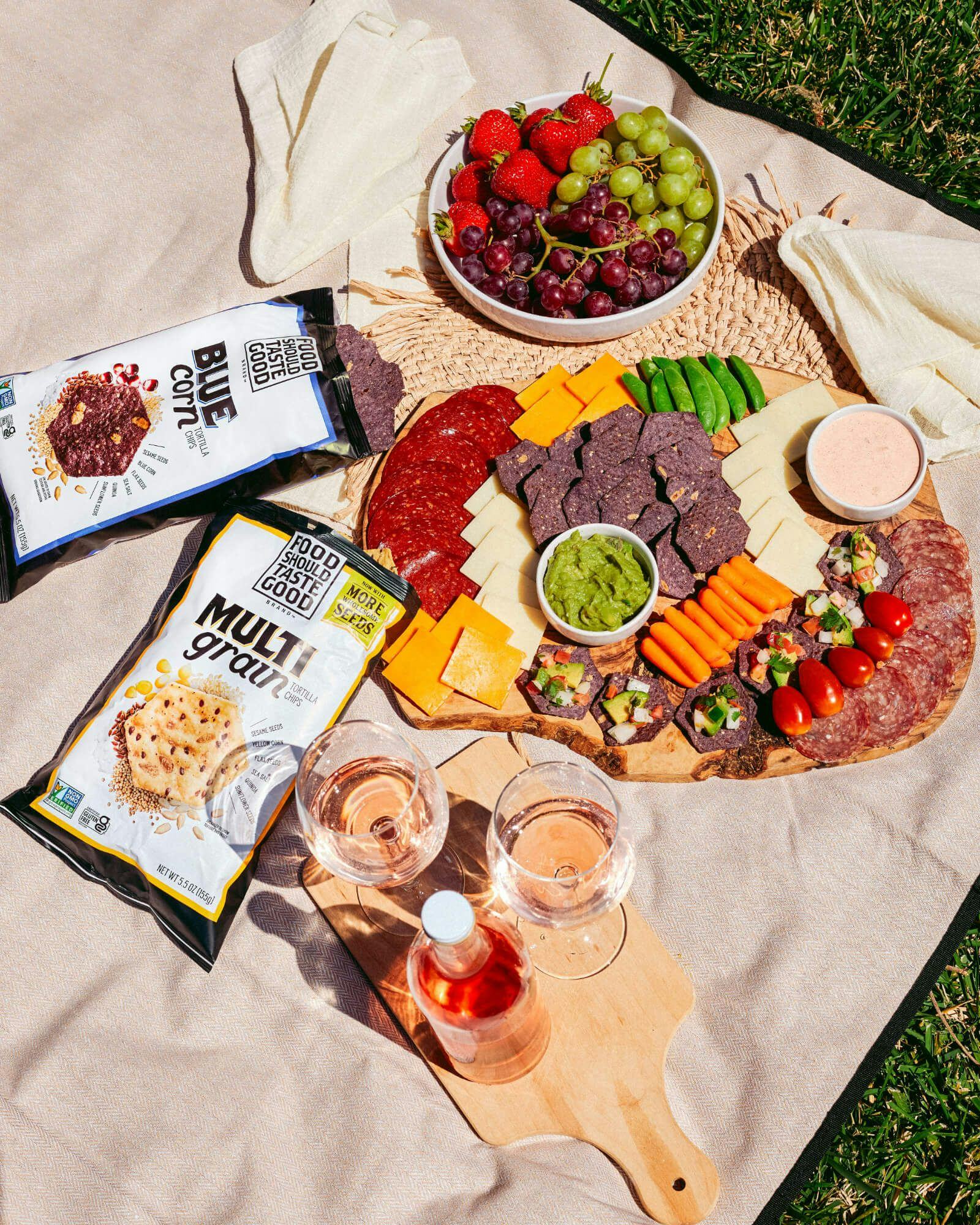 Bags of chips, a charceuterie board, and beverages laid out on a picnic mat.