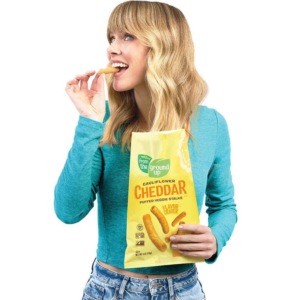 Smiling woman holding a From the Ground Up chip bag in one hand, a chip in the other.